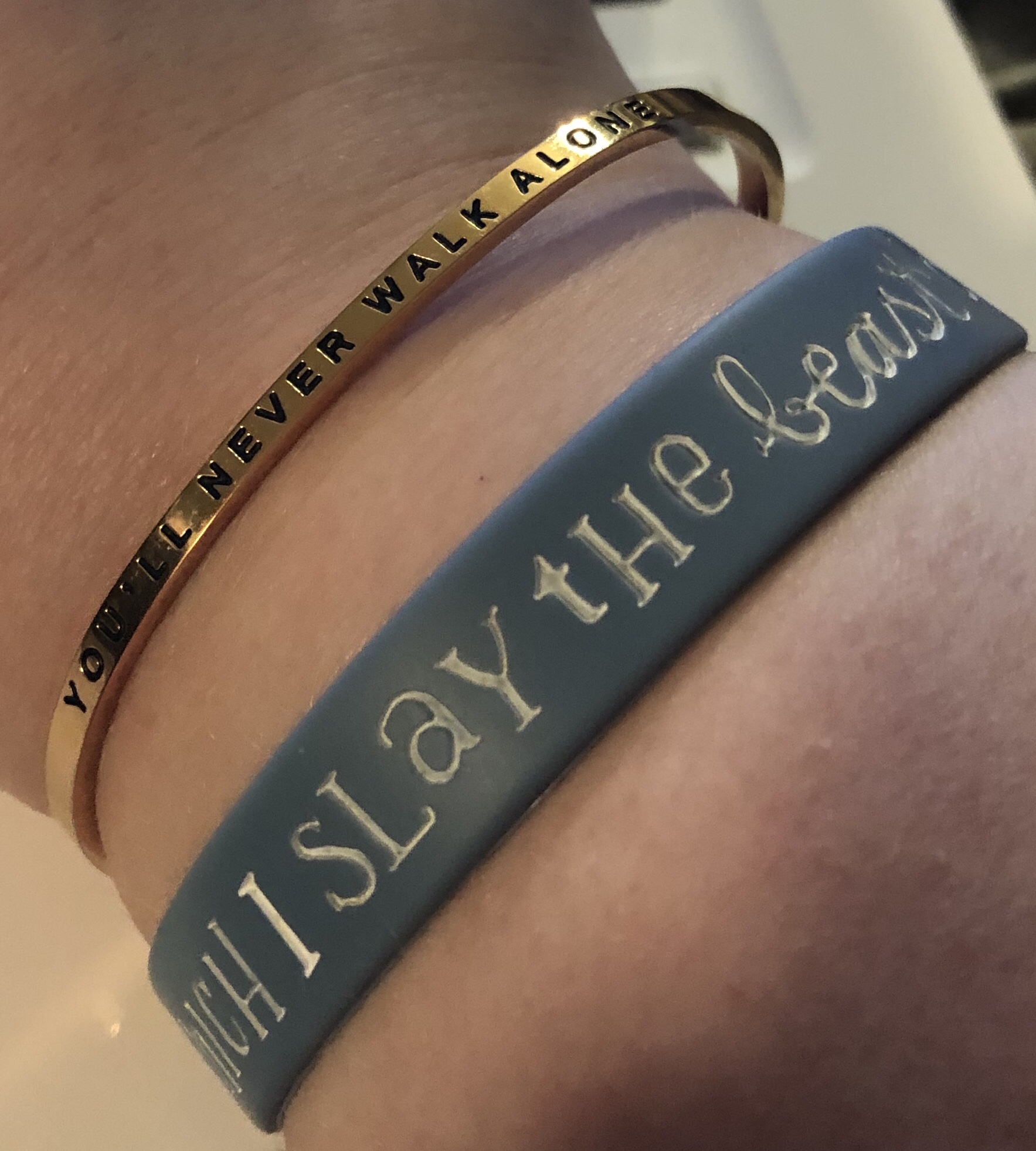 Photo of two bracelets on a wrist. The first says "You never walk alone" and the second shows the text "I slay the beast".